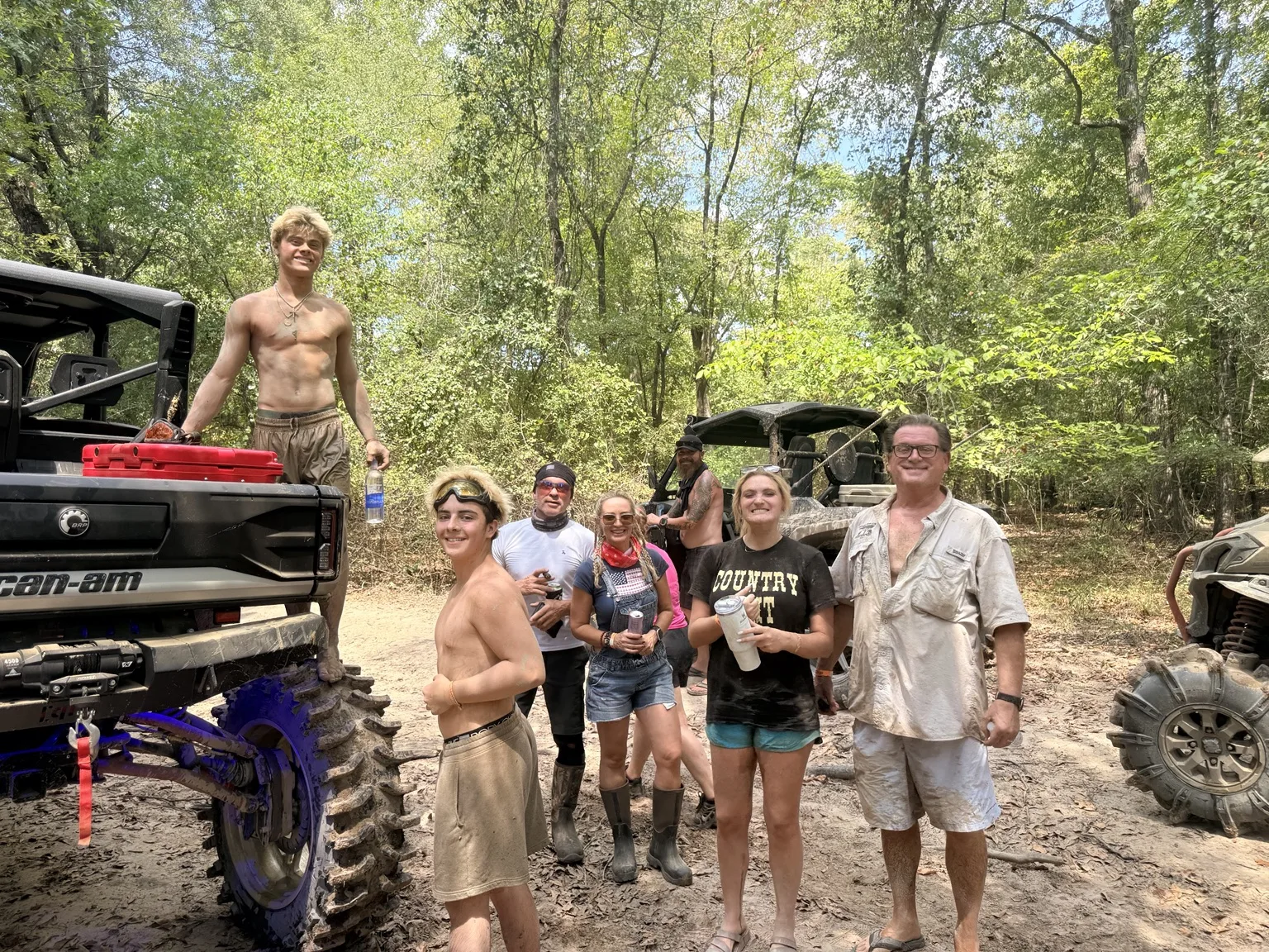 Friends smiling beside muddy Can-Am side-by-sides on a sunlit wooded trail