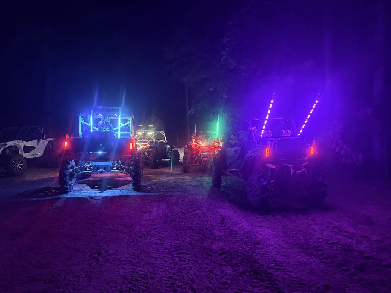 Off-road side-by-sides gathered on a wooded trail at night, glowing with blue, green, and purple lights