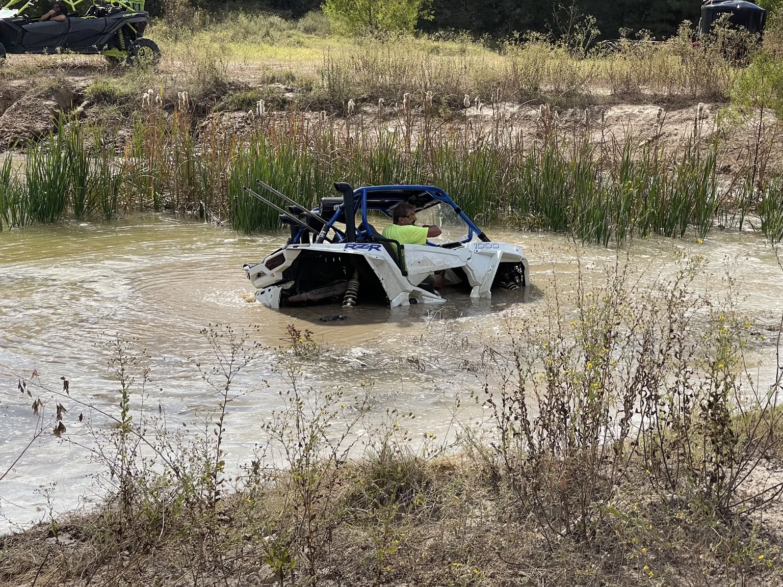 A rider in a white Polaris RZR crossing muddy water with other side-by-sides nearby